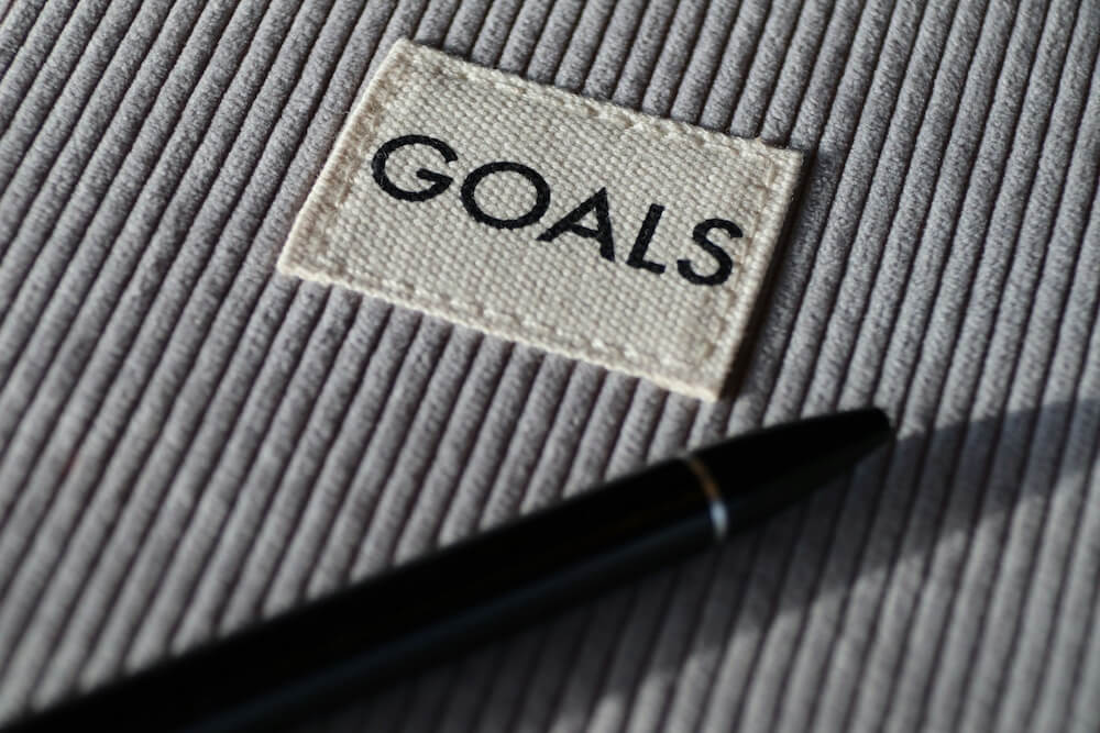 close-up view of gray corduroy fabric with a small white canvas patch sewn on with white thread. the patch reads "GOALS" in all-caps black letters