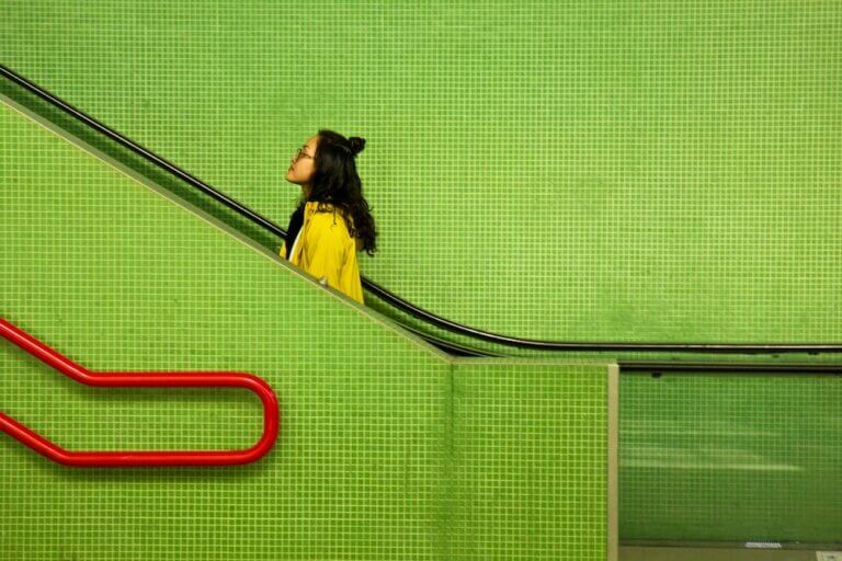 Bright green square tiles cover the walls and side of an escalator in a subway station. A young woman in a yellow sweater stands near the bottom of the escalator, looking up