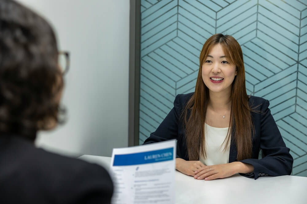 A young Asian woman with long light-brown hair and curtain bangs wears a navy blazer and white blouse. She sits at a white conference table with hands resting on the table, across from someone with short wavy brown hair and glasses who is holding a resume. It looks like a job interview; the young woman looks eager and hopeful as she smiles at the interviewer.
