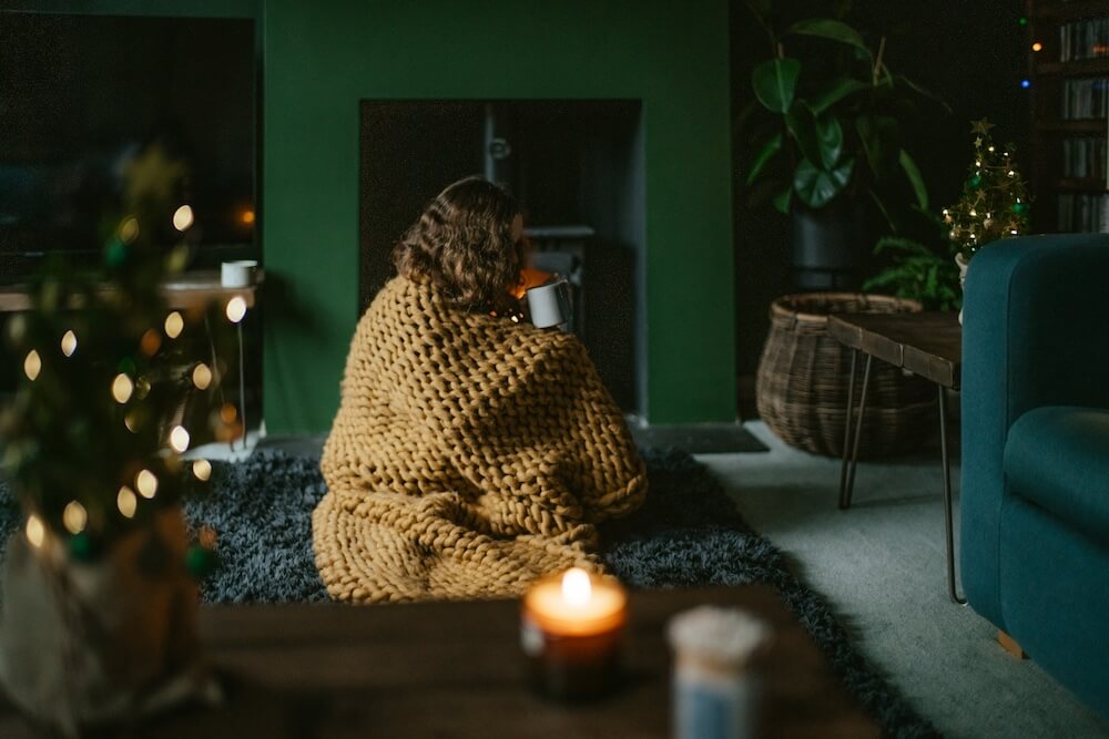 Person with curly hair sits on the floor, wrapped in a chunky brown knit blanket and holding a mug up to their lips. Candles are lit and the green living room is dim, creating a cozy ambience