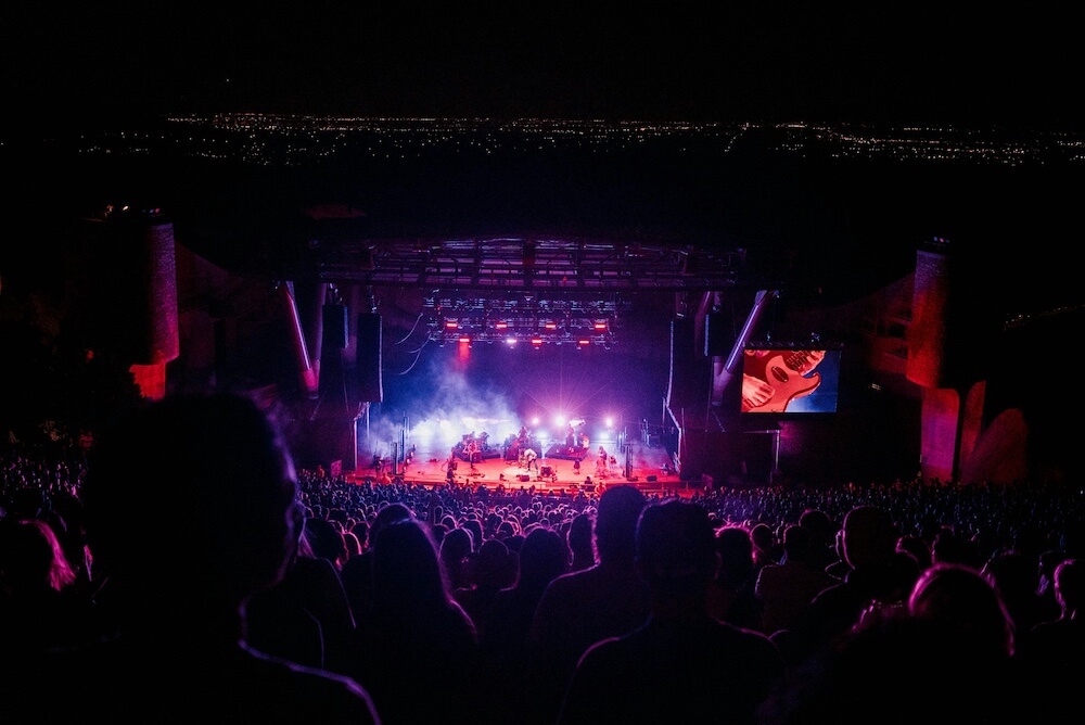 The view of a concert arena from far back seats. The stage lights are purple and red and we see the outline of the audience illuminated by the lights. 