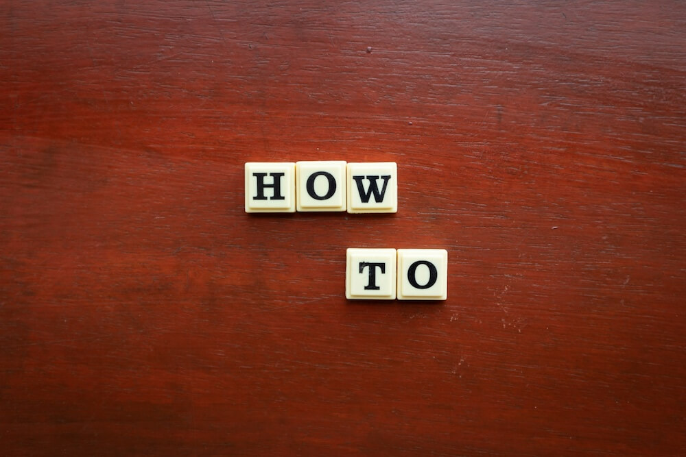 A mahogany wood table top with white blocks, each with a large capital letter in black ink. Together, the blocks spell "HOW TO"