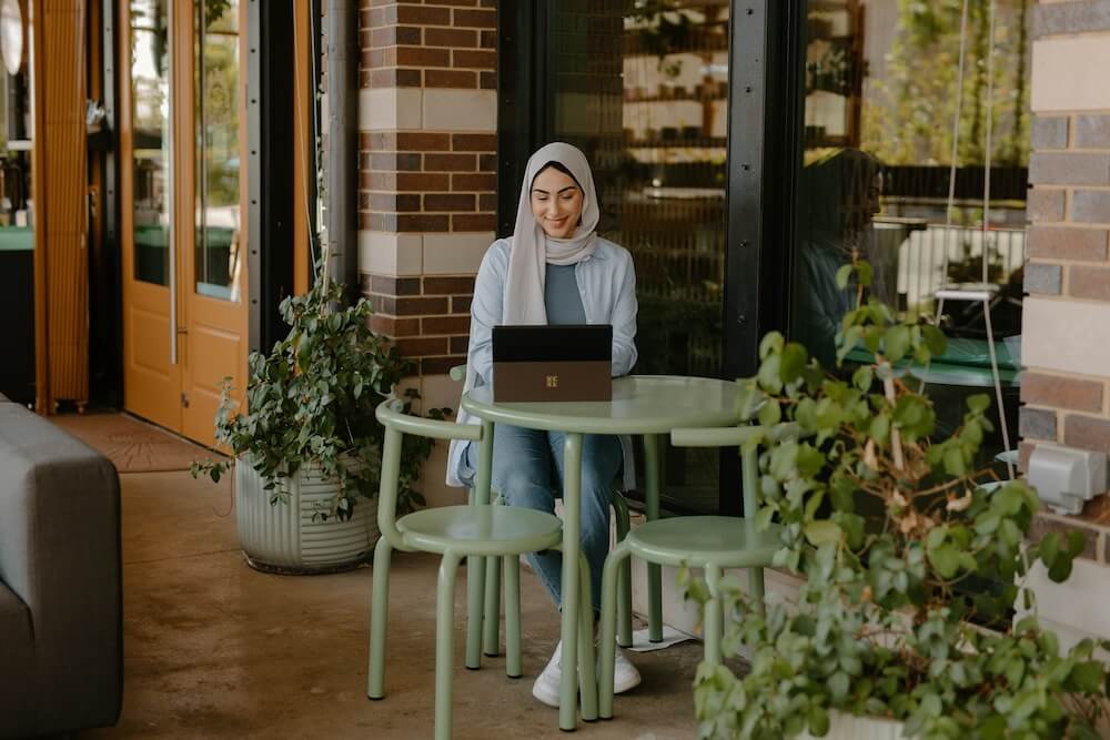 A young woman with beige religious head scarf wears a blue cardigan, blue shirt, jeans, and white shoes, sitting at a light green table outside of a coffee shop. She's got a tablet on the table in front of her and looks down at the screen, smiling, as though on a video conference — possibly with a career transition coach, mentor, or colleague.