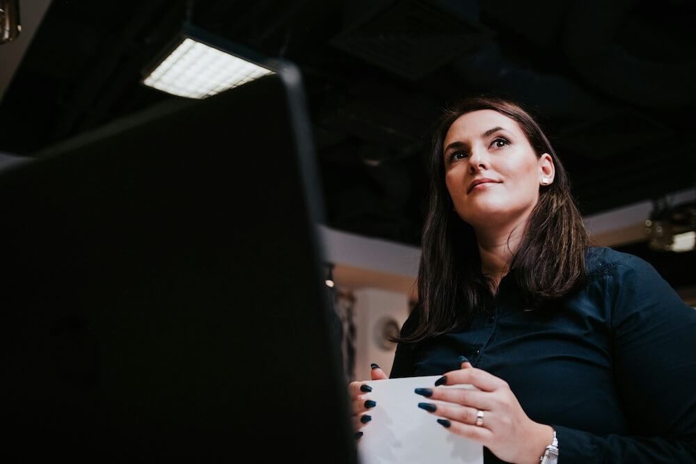 woman with long brown hair just below shoulder wears a navy button-down shirt and has long black fingernails. she holds some paper and stands in front of a desktop screen, but is looking off to the side in a dark office environment, with a slight smile