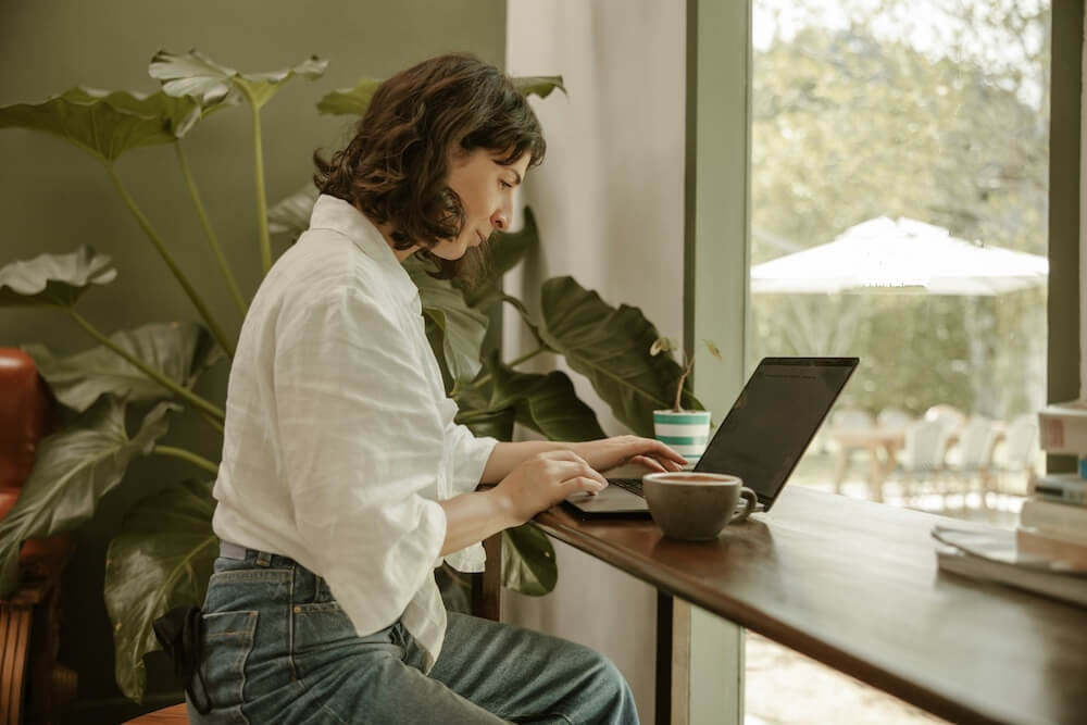 A feminine-presenting person with bob-length wavy dark brown hair and bangs sits on a stool at a window countertop, presumably in a coffee shop. She wears a white collared shirt and light blue jeans. She's looking at her laptop screen and getting ready to click on something. There's a mug on the counter next to her. 