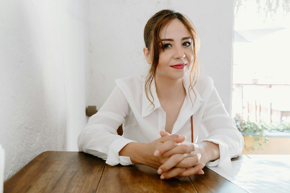 A young woman with dyed reddish-brown hair sits at a wooden table with hands clasped. The room is brightly lit by the sun, with white walls and  a view of the outside. She wears a white button down shirt, with red lipstick and light pink nail polish. She tilts her head slightly and looks at the camera with a subtle smile.