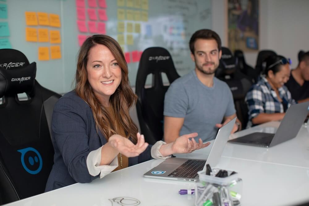 A conference room with a white table and whiteboard covered in colorful sticky notes. A 30-something white woman with long light brown hair sits at the table, wearing a blue blazer with sleeves rolled up. She gestures with both hands out and has a slight smile as though explaining or listening to someone across the table. A white male colleague with gray t-shirt, brown hair, and beard sits next to her and listens. 