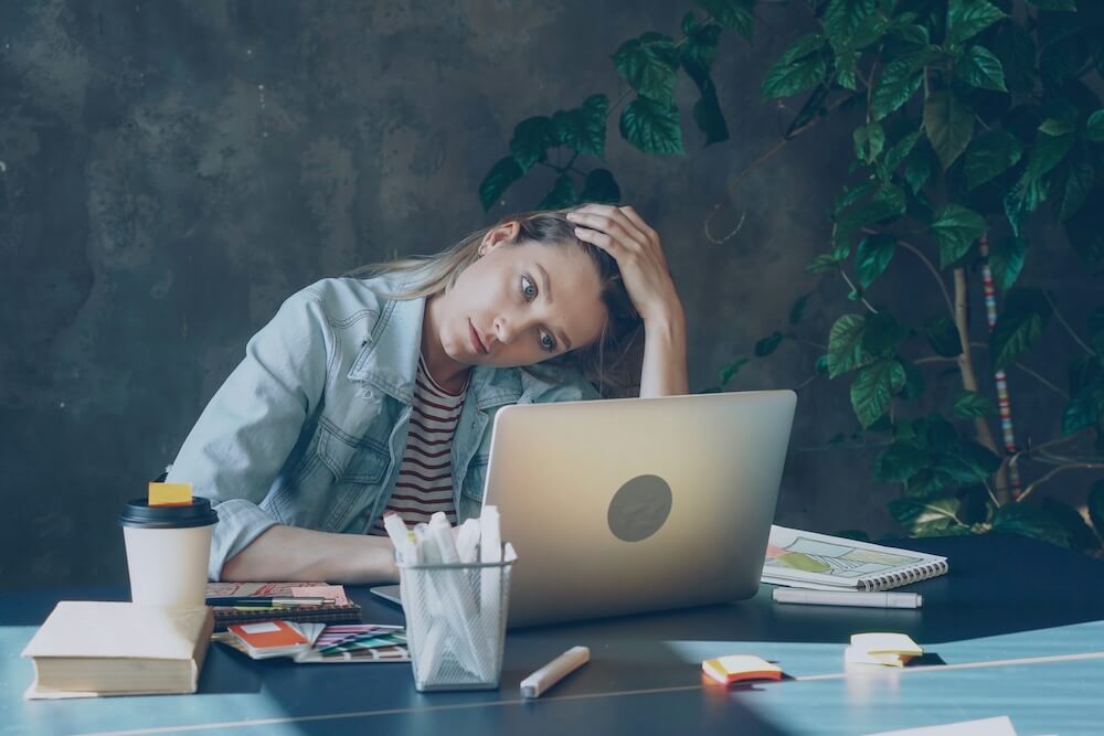 A white woman with brown hair pulled into ponytail leans on a desk in front of her, with head tilted to the side and one hand covering the top of her head. She looks exhausted as she stares at her laptop screen. 