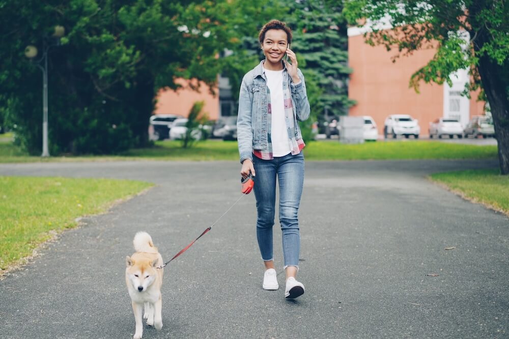 A young black woman with short hair wears a denim jacket, cropped skinny jeans, and white sneakers. She holds her phone up to her ear with one hand, and holds the red handle of a retractable leash in the other; her Shiba Inu dog walks on the pavement in front of her. They're in a city park.
