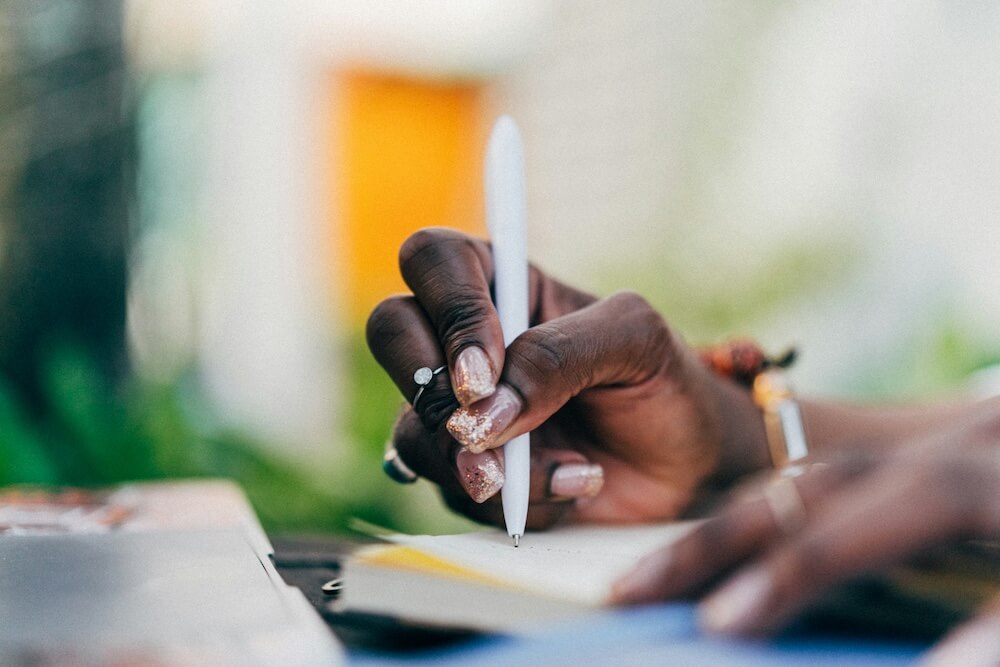A close-up of a Black woman's hand holding a white pen and writing. She has a square-tipped clear manicure with gold glitter near the ends of each nail, and a simple silver ring with a round diamond worn on the middle knuckle of her middle finger.