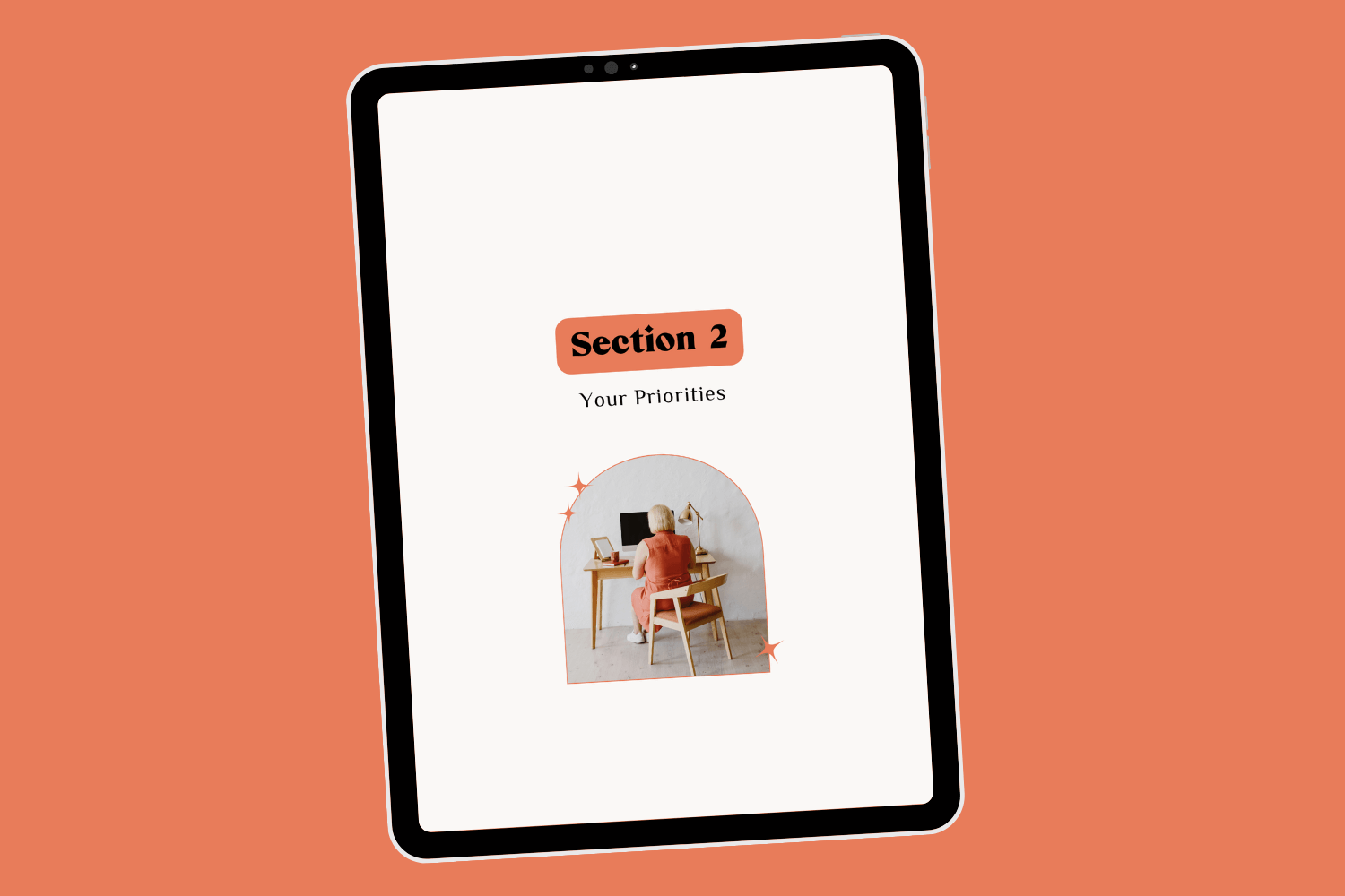 Tablet with page of ebook career transition guide Career Clarity Workbook by Lucy Todd Coaching. The title of the page is "Section 2: Your Priorities" featuring a photo of a young white blonde woman in an orange dress facing away from us, sitting at a desk working on her computer in a naturally-lit white home office space