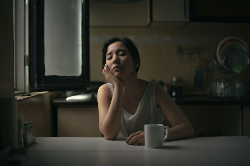 An Asian woman sits in a dark kitchen with the lights off, slightly illuminated by a nearby window. She rests one elbow on a table and props her head up in that hand. The other hand rests on the table, near a white mug. Her eyes are closed and she looks exhausted — not just fatigued, but like she's tired of something in her life.
