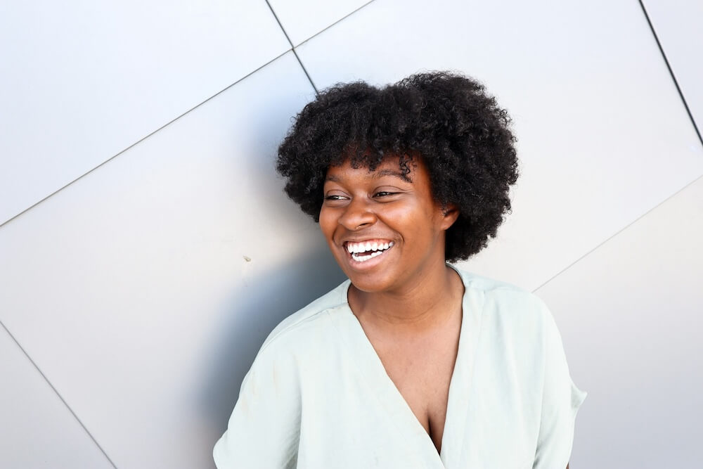 A young Black feminine-presenting person with natural short hair looks off to one side with a big smile, as though in the middle of laughing. She wears a white loose-fitting shirt with a low v-neck and stands in front of a white tiled wall.