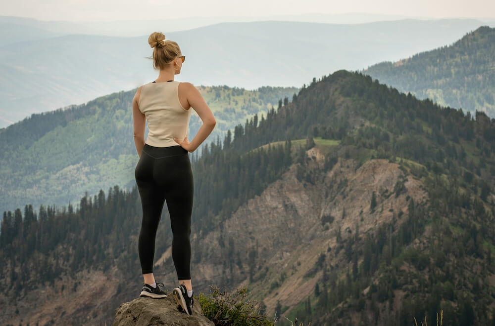 blonde woman in sleeveless cream-colored athletic top, black leggings, and black and white athletic shoes wears her hair in a bun and stands on top of a rocky peak, looking out at the evergreen-covered hills in front of her. 