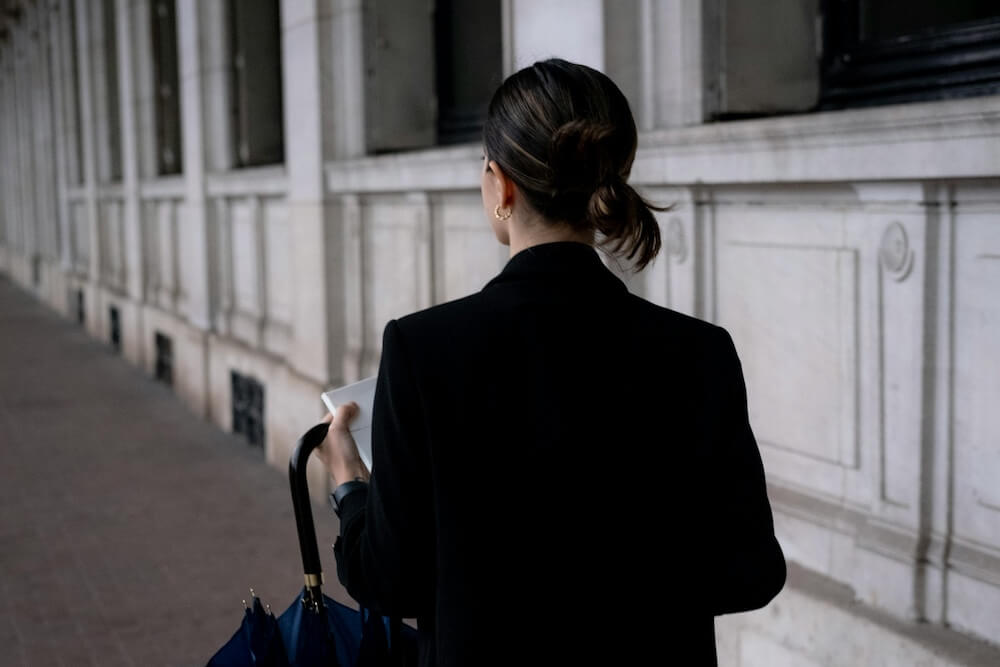 A young woman with black hair and blonde highlights has back turned to the camera, walking away down a city street. She wears a black jacket and gold hoop earrings and holds a notebook and umbrella in her left hand. 