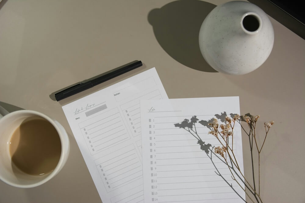 A beige desktop with a coffee mug, round white ceramic vase with slim single-stem opening, and a stack of notebooks with checklists. Some dried flower stems rest artfully near the checklists.