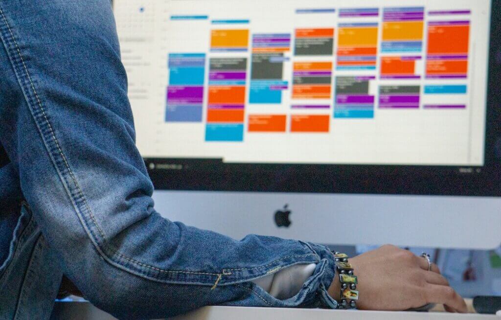 the arm of a woman wearing a long-sleeve denim jacket and resting her hand on a mouse or trackpad atop a desk. We can see part of a screen on a Mac computer display, with an out-of-focus view of Google Calendar showing a packed schedule with multicolored event boxes.