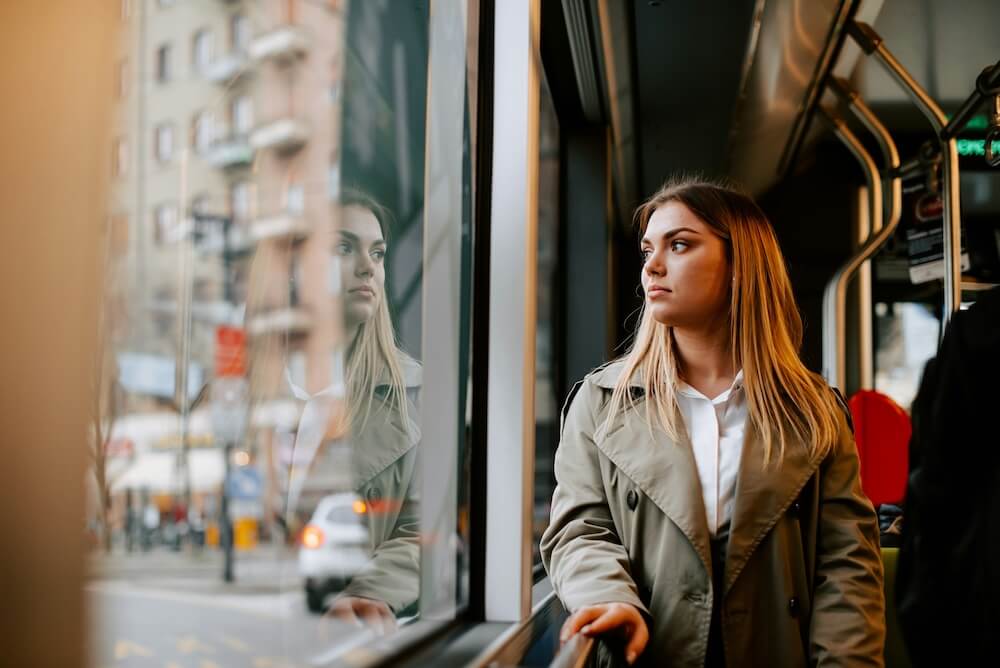 A corporate millennial woman, white with dyed blonde hair that's closer to brown at the roots, rides a modern bus or streetcar and stares out the window. She wears a khaki trenchcoat and white button-down shirt, holds a railing that runs parallel to the windows, and we can see her reflection in the window as though she's looking at herself. A tall brick building and car with brake lights on are visible, but out of focus, through the window.