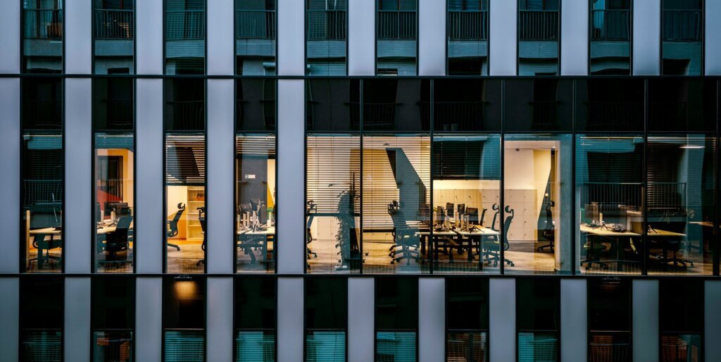 external view of an office building, showing several conference rooms and offices through the windows. the overall mood is bleak; shades are drawn to varying heights and most of the lights on the floors above and below are turned off. overall, it's a bleak corporate office