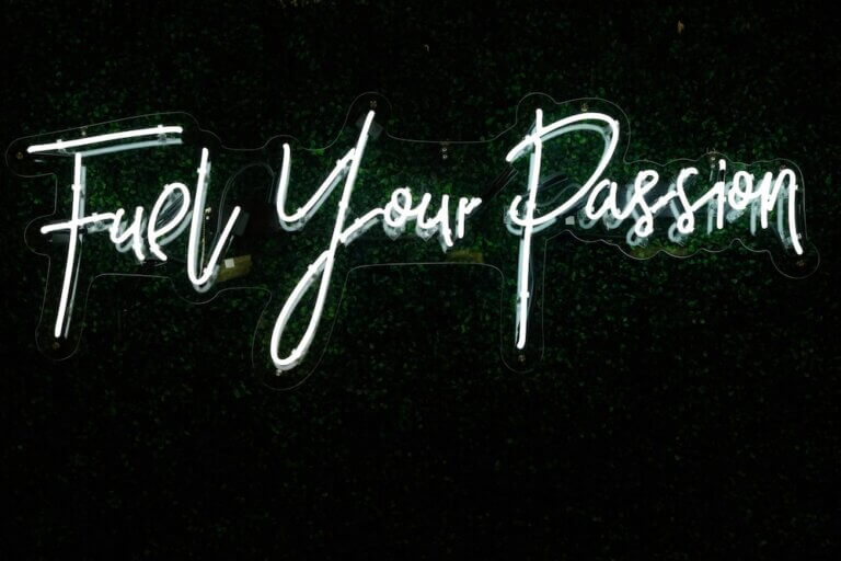A white neon sign over a dark topiary background. In a playful script font, the sign reads "Fuel Your Passion."
