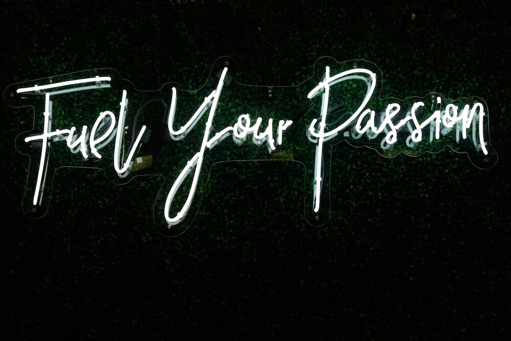 A white neon sign over a dark topiary background. In a playful script font, the sign reads "Fuel Your Passion."