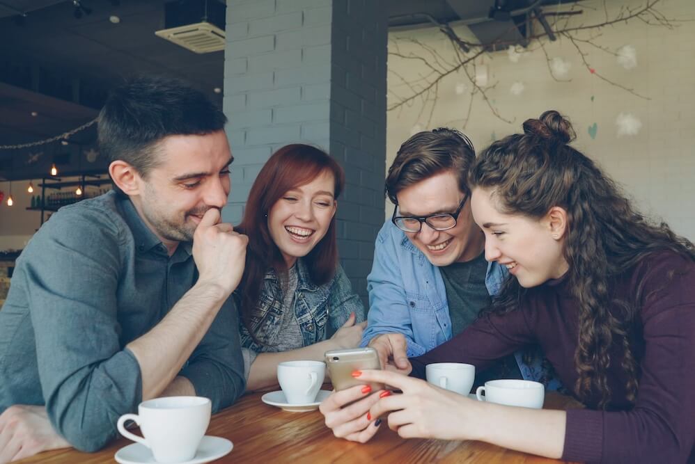 A group of coworkers, all light-skinned, two masculine-presenting and two feminine-presenting, sitting at a table in a coffee shop, They all have white ceramic mugs in front of them and are leaning in to look at a phone one of them is holding. All are interested and smiling.