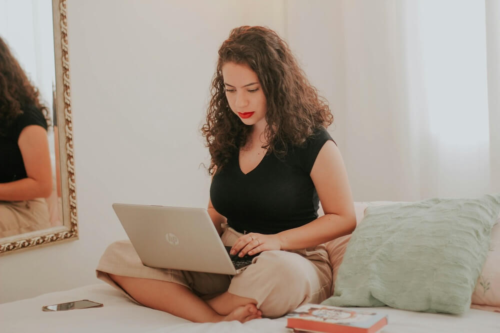 Latina woman with curly shoulder-length hair sits cross-legged on a bed, with laptop in her lap. She wears red lipstick, a black cap-sleeved t-shirt and cream-colored linen cropped pants. Her hands are on the laptop keyboard as though about to type. The room is softly lit by a window behind her, covered in a sheer white curtain.