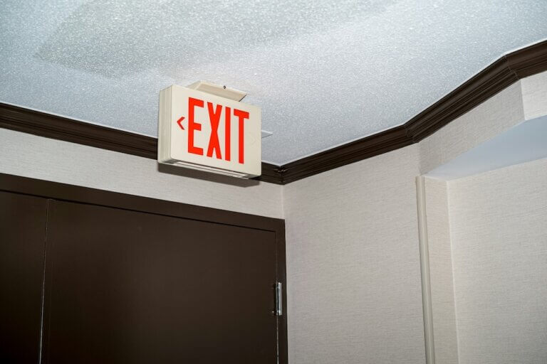 a white office ceiling with espresso brown trim and brown steel door. a cream-colored sign with red all-caps lettering reads "EXIT" with an arrow pointing to the left.