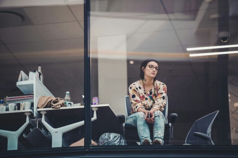 A white woman with black bob haircut and glasses sits in an office chair, turned toward the window. She looks out the window with an unfocused gaze, elbows resting on her legs and hands clasped between her knees, holding her phone. The overall mood is somber, like she's dissatisfied with her career but unsure what to do about it.