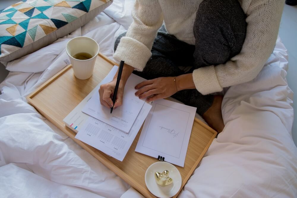 A woman sitting cross-legged on a fluffy white comforter on a bed, wearing sweatpants and a thin white sweater with cuffed sleeves. She holds a black pen and writes on a notebook that sits on top of a monthly planner, on a lap desk that rests on the bed. A white cardboard to-go cup of tea sits on the lap desk, as well as a decorative dish with a gold figurine.
