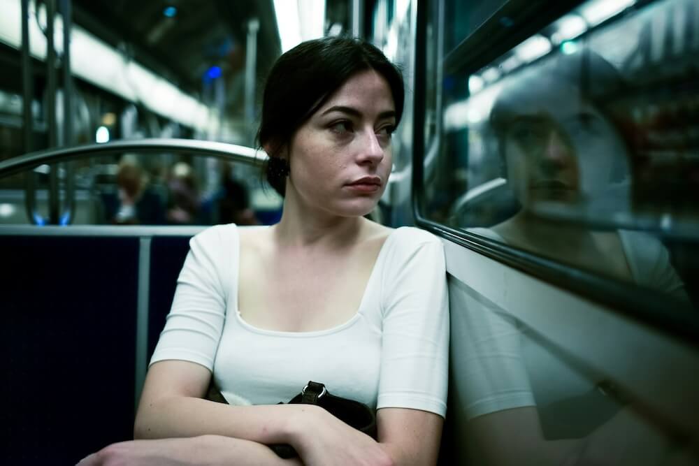 A white woman with black hair sits on a subway, staring sadly out the window. She wears a white scoop-neck top with sleeves that stop just before the elbow; her arms are folded on top of a coat or sweater that rests on her lap.