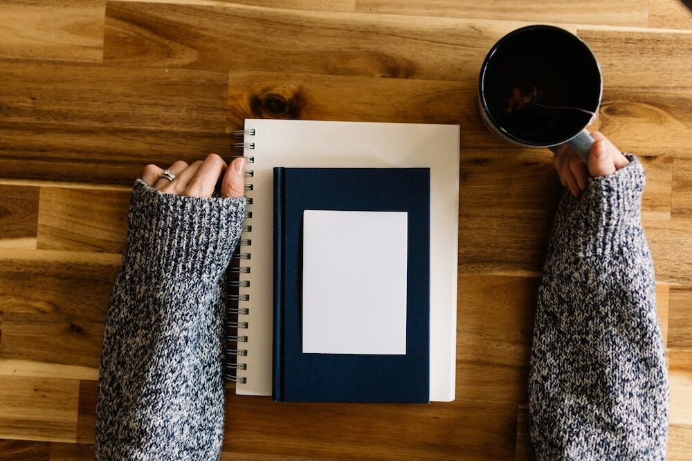 POV photo of a woman's arms resting on a wooden tabletop, wearing a dappled knit sweater and with a white-gold engagement ring and wedding ring set. She holds the handle of a mug of tea in one hand, resting the other hand with fingers gently folded on the table, thumb on the edge of a spiral-bound notebook. On top of the notebook is another smaller navy-blue notebook, and a small white notepad on top of that. It looks like the woman is getting ready to journal or self-reflect.