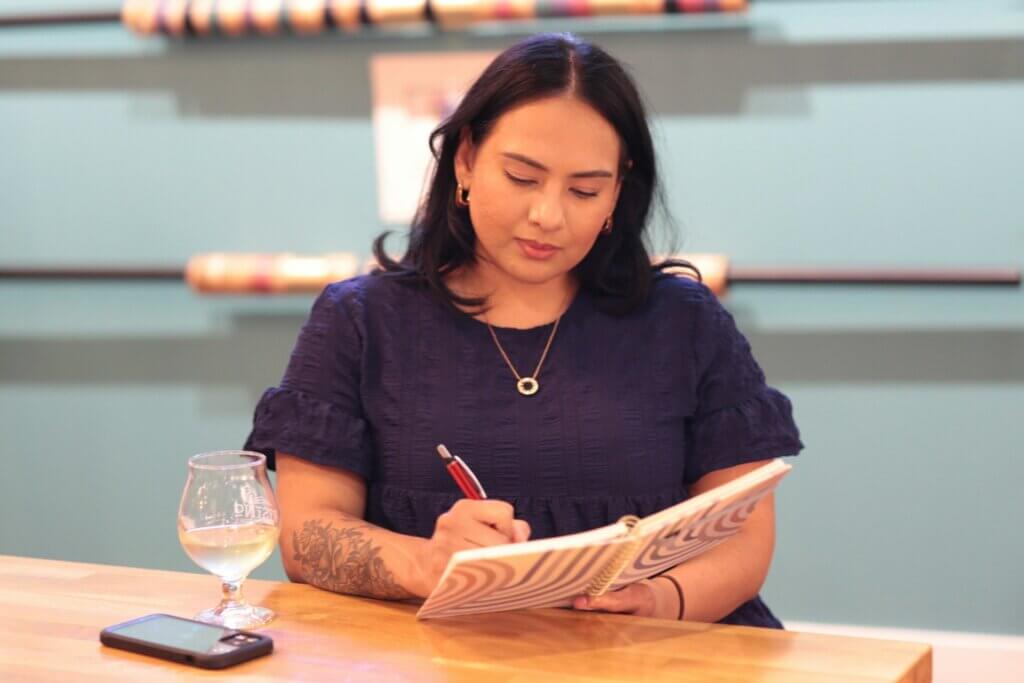 A young woman of color with black hair parted down the middle and intricate tattoo on her forearm sits in a brewery setting, a fancy beer glass on the table in front of her. She wears a frilled navy-blue shirt, gold hoop earrings, and gold necklace with circular pendant. She holds a red pen and is writing in a spiral-bound notebook, concentrating and deep in thought.