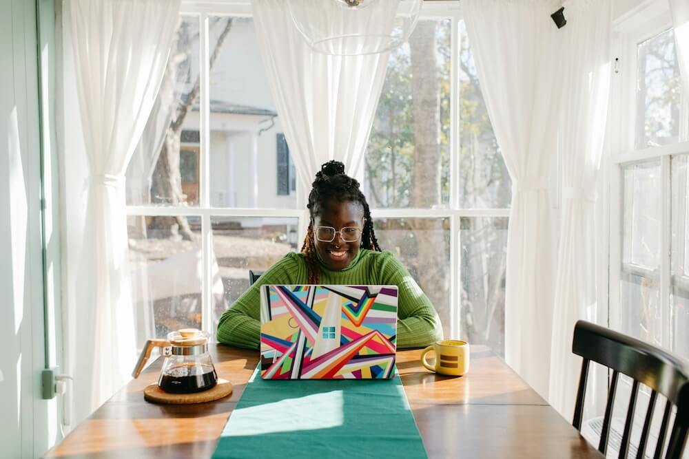 A Black woman sits at a long wooden dining table in a bright dining room. She wears a green sweater, gold rectangular glasses, and her hair in braids. She rests her elbows on the table and smiles at her laptop screen, which has a colorful geometric case. 