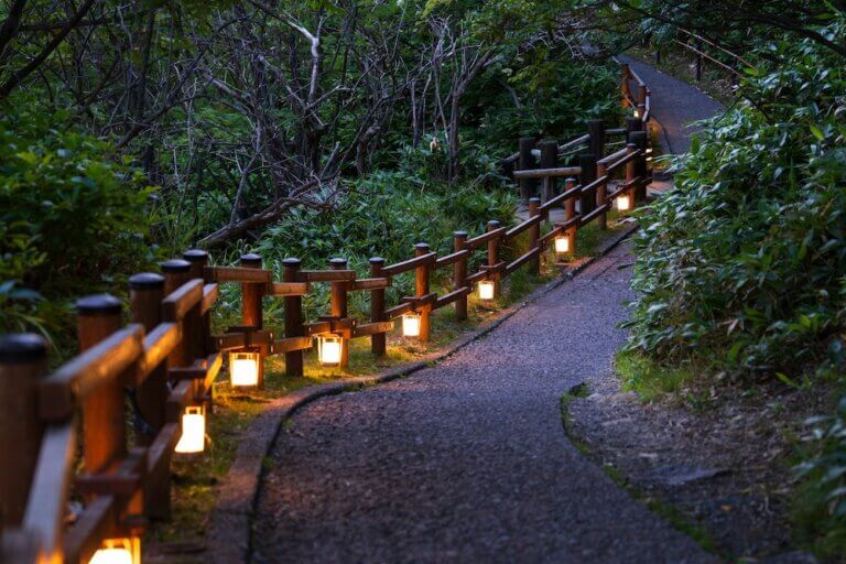A gravel pathway in dim light, illuminated by candles alongside a rustic short wooden fence. Lush greenery on either side of the path
