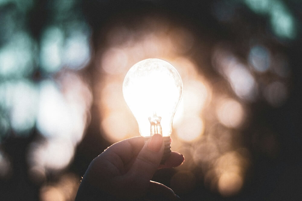 A person with orange nail polish holds a lightbulb up in front of rays of evening or morning sunlight, making it look like the lightbulb is illuminated.