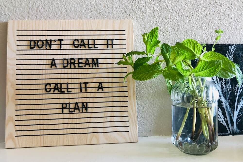 A white desktop with a mason jar full of mint and a light wood letter board. The black block letters read "DON'T CALL IT A DREAM / CALL IT A PLAN"