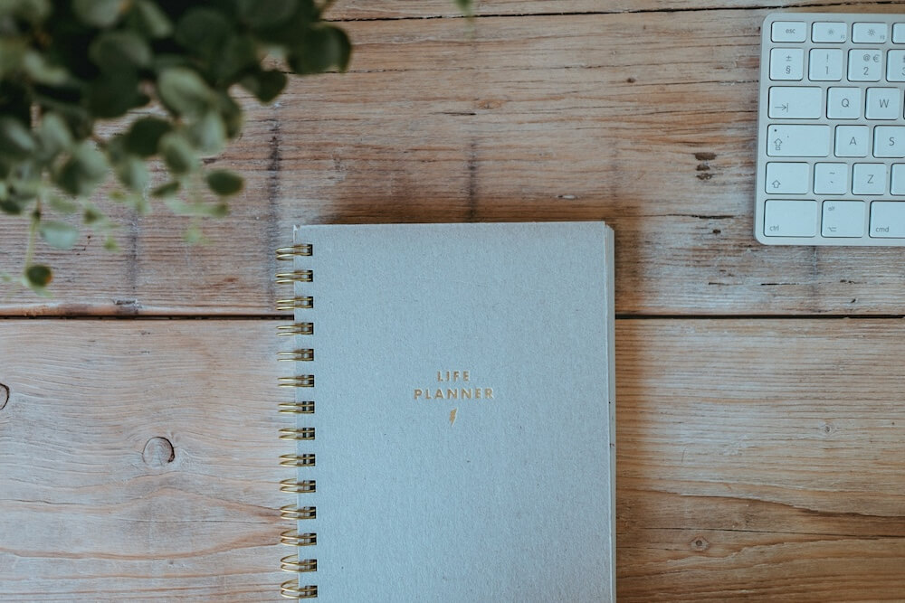 An unfinished wooden tabletop with a seam between boards, like a picnic table but indoors. There's a green houseplant and white Apple keyboard on either side of the image, and in the center is a spiral-bound notebook with the words "LIFE PLANNER" written in gold.