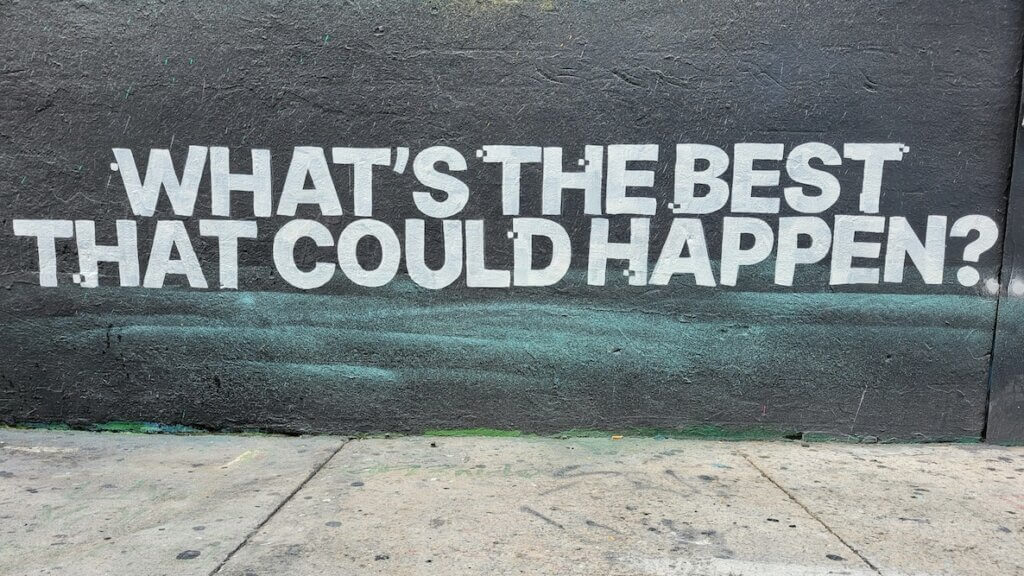 The base of a black exterior wall and a gray sidewalk. In white block all-caps letters stenciled on the wall is the question, "WHAT'S THE BEST THAT COULD HAPPEN?"
