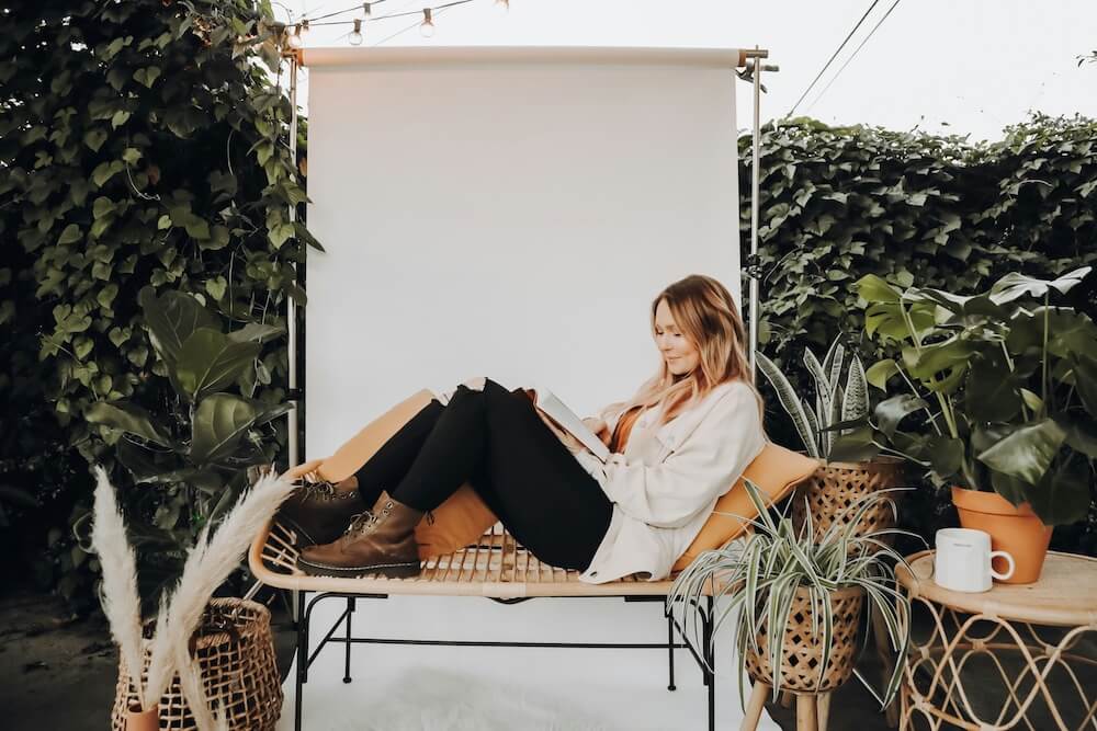 A white woman with light brown/blonde balayage hair reclines on a wicker loveseat in a garden patio with potted plants and an ivy-covered wall. She wears a white sweater, black skinny jeans, and brown Doc Marten ankle boots. She's reading or writing in a book and smiling.
