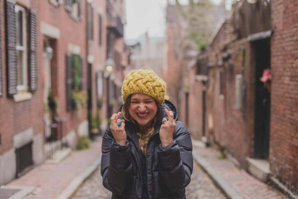 A young white woman in oversized yellow beanie and black puffy winter jacket stands in an alley lined with brick buildings. She crosses her fingers tightly and scrunches her face with a smile, wishing for something she really wants.