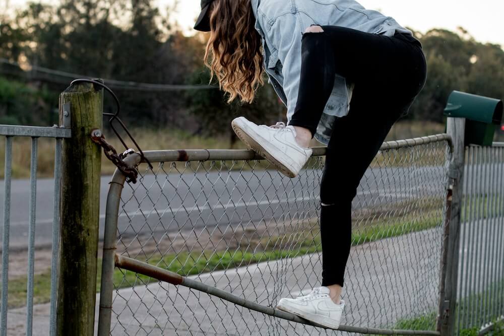 A white feminine-presenting person with long brown wavy hair wears a denim shirt, black skinny jeans, and white sneakers. She climbs a low chain-link gate, one foot on a railing and the other on the top. She's about to jump over to the other side.