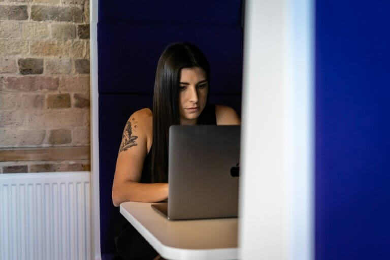 A white millennial woman with long, straight, dark brown hair sits in an office pod with blue walls and a white table. She wears a black tank top and has a black-and-white tattoo on her upper arm. She stares somewhat blankly at the laptop screen in front of her, thinking.