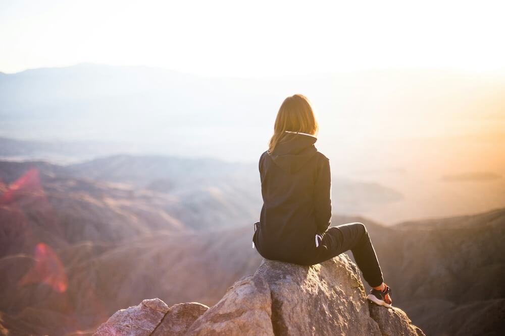A white woman with light brown hair and blonde highlights sits with her back to the camera atop a mountain peak. Other hills and a lake are visible, but out of focus. She wears a black hooded jacket, black skinny joggers, and black and red athletic shoes. Her head is lifted up slightly, like she's taking in the view.