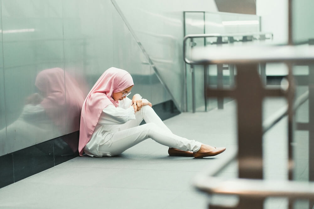 A woman in pink hijab, white skinny jeans, and beige flats sits on the floor of a modern office building, leaning against the wall of a staircase or escalator. She rests her folded arms against one raised knee and looks down, seeming sad and deep in thought. 
