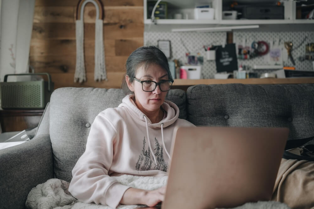 A white millennial woman in light pink hoodie and black glasses sits underneath a fuzzy white blanket on a gray couch in a home office or studio. Her hair is messily tied back and she looks tired, like she's working first thing in the morning or just disengaged at work. There's a laptop on her lap and she's typing and looking at the screen.