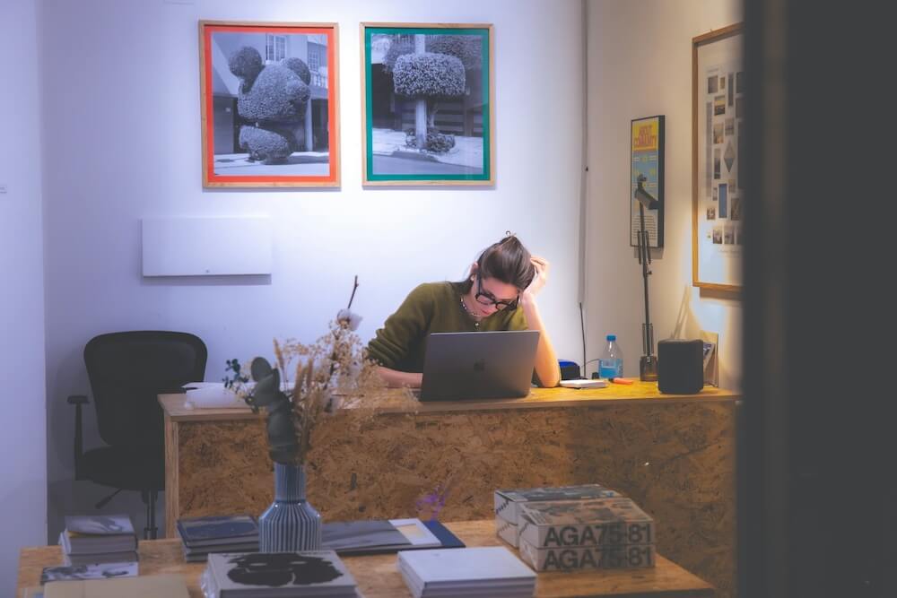 A white brunette woman in a green sweater rests an elbow on her wooden desk and leans her head on her hand. She looks tired or upset, staring at her laptop. The office is dimly lit with only a lamp, making it look like an early morning or late night workday.