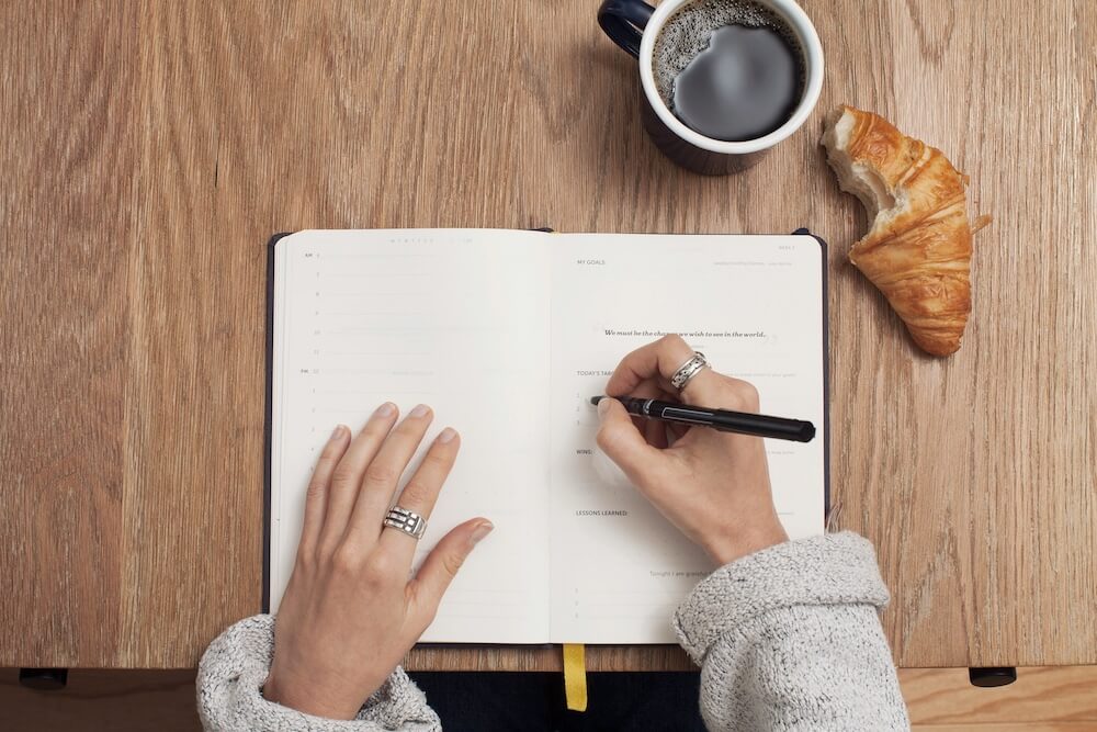 The hands of a white feminine-presenting person who wears a silver ring and a light-gray sweater cuffed slightly at the sleeves. They hold a black pen in one hand and are writing in a notebook with specific boxes allotted for goals and other topics. The notebook sits atop a wooden desk, alongside a mug of black coffee and a croissant with a bite taken out of it.