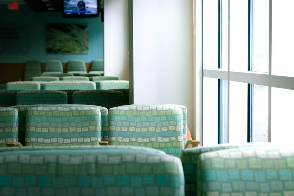 An empty hospital waiting room with rows of patterned green chairs and a large floor-to-ceiling window
