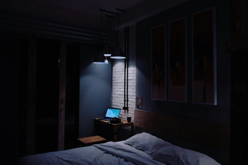 A dark bedroom with bed and nightstand, lit only by decorative pendant lights over the nightstand. A laptop sits open on the nightstand, as though someone has been working long hours late into the night.